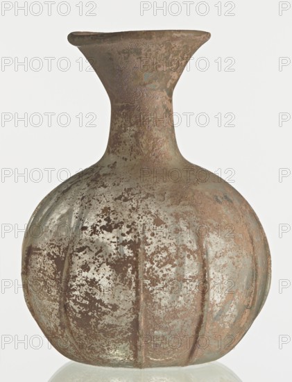 Flask; body in the shape of a flattened sphere, decorated with ribs; maker, Syria, 2nd–3rd century AD. Type: Isings form 101? Transparent, colourless, iridescent glass. Slightly flattened spherical body decorated with vertical thin, uneven ribs; short cylindrical neck terminating in a funnel-shaped spout with a thickened and rounded edge; free-blown. Outer diameter of the spout: 3.80 cm. Height: 8.80 cm. Body diameter: 8 cm. Bottom diameter: 3.40 cm. Vessel; weight 0.030 kg., A rounded, bulbous vessel with a narrow neck and flared rim fills the frame, showing a weathered, pitted surface texture. The object displays a mix of brown, tan, muted green, and pale gray tones with mottled patches and speckled corrosion-like marks across its body. The vessel has subtle vertical ridges on its lower half and an uneven, rustic finish that gives it an aged appearance. The background is a plain light color that contrasts with the object.