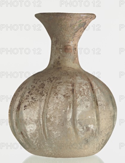 Flask; body in the shape of a flattened sphere, decorated with ribs; maker, Syria, 2nd–3rd century AD. Transparent, colourless, iridescent glass. Slightly flattened spherical body decorated with vertical thin, uneven ribs; short cylindrical neck terminating in a funnel-shaped spout with a thickened and rounded edge; free-blown. Outer diameter of the spout: 3.80 cm. Height: 8.80 cm. Body diameter: 8 cm. Bottom diameter: 3.40 cm. Type: Isings form 101? Weight: 0.030 kg., A rounded glass vessel with a bulbous body and a narrow flaring neck, covered in a weathered, pitted surface showing mottled tones of pale green, muted brown, beige, and faint iridescent sheen, with vertical ridges on the body and areas of darker brown speckling and encrustation.