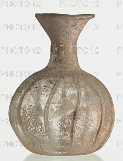 Flask; body in the shape of a flattened sphere, decorated with ribs;, Syria; 2nd–3rd century AD. Transparent, colourless, iridescent glass. Slightly flattened spherical body decorated with vertical thin, uneven ribs; short cylindrical neck terminating in a funnel-shaped spout with a thickened and rounded edge; free-blown. Outer diameter of the spout: 3.80 cm. Height: 8.80 cm. Body diameter: 8 cm. Bottom diameter: 3.40 cm. Type: Isings form 101? Vessel; weight 0.030 kg., A round, bulbous glass vessel with a flared neck and narrow shoulder, showing a textured, mottled surface; the glass appears cloudy and weathered with patches of rough encrustation and smooth areas, and features shallow vertical ridges along the body; visible colors include pale green, muted brown, gray, and hints of beige.