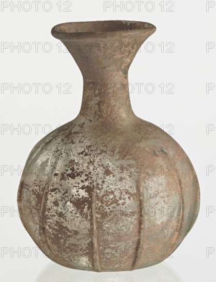 Flask; body in the shape of a flattened sphere, decorated with ribs;, Syria, 2nd–3rd century AD. Transparent, colourless, iridescent glass. Slightly flattened spherical body decorated with vertical thin, uneven ribs; short cylindrical neck terminating in a funnel-shaped spout with a thickened and rounded edge; free-blown. Outer diameter of the spout: 3.80 cm. Height: 8.80 cm. Body diameter: 8 cm. Bottom diameter: 3.40 cm. Type: Isings form 101? Weight: 0.030 kg., A rounded vessel with a narrow neck and flared rim, showing a textured surface with mottled areas and patches; the body has vertical ridges and an uneven, pitted appearance. The visible colors include muted brown, rusty orange, pale green, and gray, with darker brown speckling across the surface. The rim interior appears darker, and the overall finish looks weathered and corroded.