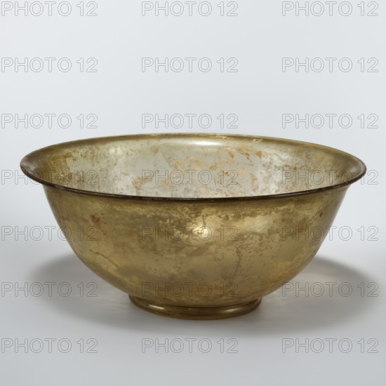 Bowl, bowl with a ring-shaped foot, walls that flare evenly upwards and a flared rim; inside the bowl the bottom is bulging. 1st–2nd century AD, Syria; vessel. Height 4.8 cm, width 12.8 cm; diameter 4.8 cm foot, diameter 12.2 cm rim., A single shallow metal bowl with a rounded base and flared rim, showing a mottled surface of metallic gold and pale greenish-silver tones with patches of darker tarnish and subtle brownish spots; the interior reflects lighter silvery and gold hues while the exterior displays more muted gold and brown variations, all set against a soft pale gray background.
