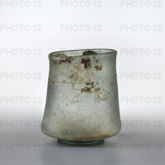 Mug with walls widely flared at the bottom above the ring-shaped foot, slightly convex and forming the bottom, and slightly tapering at the sides towards the top and ending with a thickened rim. A thin thread runs along the walls below the rim. 2nd–3rd century. Mug; vessel. Height 14.9 cm; diameter 8.9 cm; foot diameter 6.4 cm., A roughly conical vessel with a slightly flared rim sits on a flat surface; its exterior shows a weathered, matte texture with patches and speckles of brown and darker green over a predominant pale green and bluish-green base, and there are areas of lighter, almost whitish wear and subtle warm beige discoloration near the bottom.