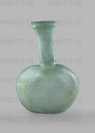 Flask with a spherical body and a high, cylindrical neck ending in a wide, straight-cut rim. The neck is marked by a recess and is decorated with a ring under the rim. Concave bottom with rounded edges. The neck is set obliquely to the body. Translucent, green glass. Spherical body, cylindrical neck slightly flaring upwards, straight rim with rounded edge, heat-worked. Neck narrowed at the bottom; free blowing. Type: Isings form 104B, 3rd–4th century, Phoenicia; vessel. Height 18.4 cm; width 13.2 cm; rim diameter 4.9 cm; foot diameter 6 cm; maximum diameter 11.9 cm., A rounded glass bottle with a bulbous body and a long narrow neck topped by a flared rim sits against a plain neutral background; the bottle is translucent with a pale greenish-blue color and shows subtle variations in tone with areas of slightly darker green, faint cloudy patches, and small speckles and weathered marks on its surface.