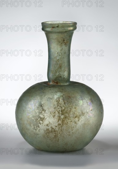 Flask with a spherical body and a high, cylindrical neck ending in a wide, straight-cut rim. The neck is marked by a recess and is decorated with a ring under the rim. Concave bottom with rounded edges. The neck is set obliquely to the body. Translucent, green glass. Spherical body, cylindrical neck slightly flaring upwards, straight rim with rounded edge, heat-worked. Neck narrowed at the bottom; free blowing. Type: Isings form 104B, 3rd–4th century, Phoenicia; vessel. Dimensions: height 18.4 cm; width 13.2 cm; rim diameter 4.9 cm; foot diameter 6 cm; maximum diameter 11.9 cm., A glass vessel with a rounded body and a long narrow neck topped by a slightly flared rim, showing an aged, weathered surface with iridescent sheens and areas of mottled patina; the visible colors include pale green, muted blue, hints of brownish rust, and subtle purple and silver reflections, all set against a neutral gray-white background.