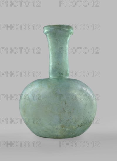 Flask with a spherical body and a high, cylindrical neck ending in a wide, straight-cut rim. The neck is marked by a recess and is decorated with a ring under the rim. Concave bottom with rounded edges. The neck is set obliquely to the body. Translucent, green glass. Spherical body, cylindrical neck slightly flaring upwards, straight rim with rounded edge, heat-worked. Neck narrowed at the bottom; free blowing. Type: Isings form 104B, 3rd–4th century, Phoenicia; vessel. Height 18.4 cm; width 13.2 cm; rim diameter 4.9 cm; foot diameter 6 cm; maximum diameter 11.9 cm., A translucent pale green glass vessel with a rounded bulbous body and a tall narrow neck that flares slightly at the rim, showing a weathered surface with some cloudy areas and patches of muted brownish discoloration, placed against a plain light gray background.