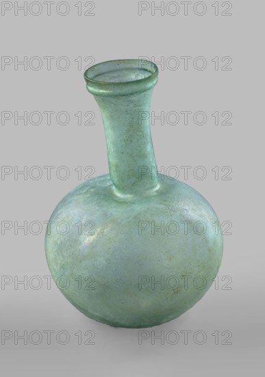 Flask with a spherical body and a high, cylindrical neck ending in a wide, straight-cut rim. The neck is marked by a recess and is decorated with a ring under the rim. Concave bottom with rounded edges. The neck is set obliquely to the body. Translucent, green glass. Spherical body, cylindrical neck slightly flaring upwards, straight rim with rounded edge, heat-worked. Neck narrowed at the bottom; free blowing. Type: Isings form 104B, 3rd–4th century, Phoenicia, vessel. Height 18.4 cm; width 13.2 cm; rim diameter 4.9 cm; foot diameter 6 cm; maximum diameter 11.9 cm. Maker: unknown., A rounded glass vessel with a bulbous body and a long narrow neck topped by a flared rim, shown against a plain neutral background; the glass surface is predominantly a pale green with variations toward bluish-green and hints of turquoise, and it shows iridescent patches and subtle cloudy areas, along with small spots and mottled discoloration in darker green and brown tones, and the interior rim and neck d...