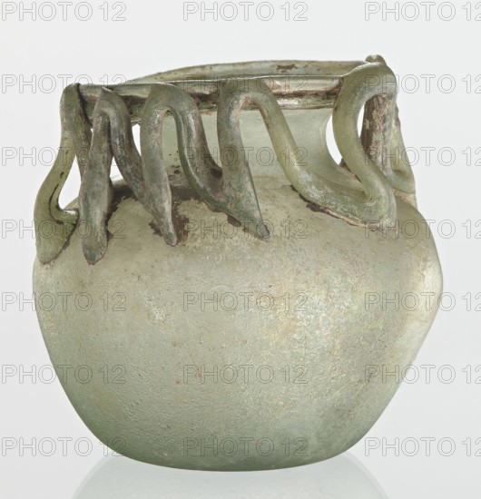 Bottle, unknown eastern workshop, 3rd century, vessel, height 7 cm, width 8.5 cm, A rounded glass vessel with a matte, slightly iridescent surface in pale green and hints of silvery sheen, topped by a flared rim from which several looping applied handles curve downward in a sinuous, ribbon-like fashion; the handles are a darker green with areas of brownish and muted coppery discoloration, and the body shows subtle mottling and weathered spots in pale green, light gray, and faint brown tones against a neutral pale background.