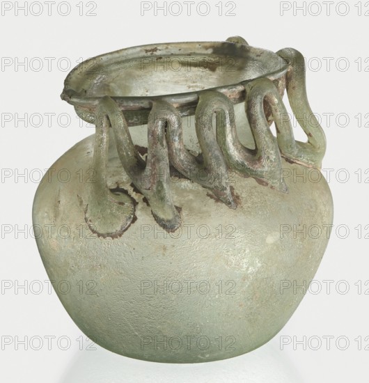 Bottle, unknown eastern workshop, Goluchów Greater Poland Voivodeship, 3rd century, vessel, height 7 cm, width 8.5 cm, A rounded glass vessel with a wide mouth and a decorative rim featuring a group of looping appendages that drape down the neck; the glass shows a weathered, textured surface with a few areas of roughness and flaking. The visible colors are pale green, muted olive, and touches of brown and gray, with lighter, almost whitish weathering in spots and subtle darker green accents along the loops. The overall shape is bulbous at the body, tapering to a narrower neck beneath the ornate rim, and the object rests on a reflective surface.