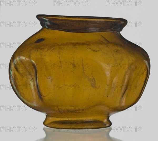 Vessel, Goluchów Greater Poland Voivodeship place of origin, vessel, height 10.2 cm, width 13.2 cm, A translucent rounded glass vessel with a short flared neck and a slightly indented foot, shown against a plain light background; the glass is amber with areas of darker brown and subtle greenish and iridescent sheen, with visible surface imperfections, small bubbles, and fine lines across the body; the shape is bulbous with gentle flattened sides and shallow indentations near the midsection, and the vessel sits reflected faintly on the surface below.