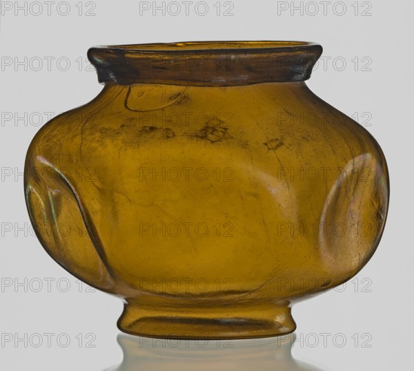 Vessel, Goluchów Greater Poland Voivodeship - place of origin, vessel, height 10.2 cm, width 13.2 cm, A rounded glass vessel with a short neck and flared rim, shown against a plain light background; the glass is an amber color with darker brown tones at the rim and base, exhibiting subtle surface irregularities and a few shallow indentations on the sides, where light creates pale highlights with faint iridescent green and purple sheens; the body has visible streaks and small surface marks, and the vessel sits on a short, slightly flared foot with a soft reflection beneath it.