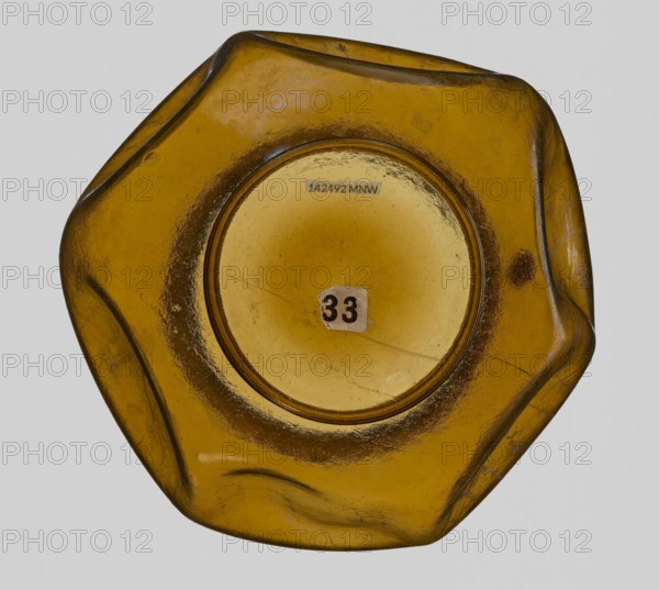 Vessel, Goluchów Greater Poland Voivodeship, vessel, height 10.2 cm, width 13.2 cm, A close-up view of a translucent amber-colored glass object with an irregular hexagonal shape and smooth, rounded edges, showing a central circular recessed area and a small worn sticker; the surface has variations in color from golden yellow to darker brown tones and shows scratches, scuffs, and a few small bubbles or imperfections.