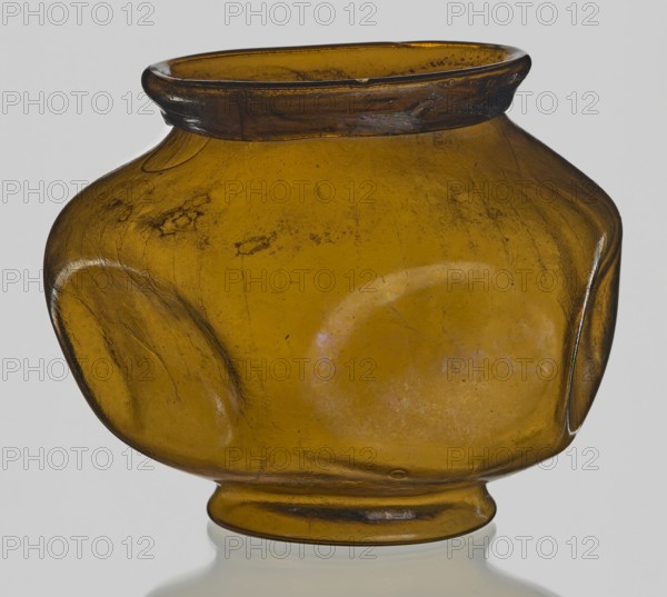 Vessel, Goluchów Greater Poland Voivodeship - place of origin, vessel, height 10.2 cm, width 13.2 cm, A rounded, bulbous glass vessel with a short neck and flared rim sits against a pale background; the glass is translucent and amber in color with variations of darker and lighter amber tones, and there are visible surface imperfections and subtle iridescent patches. The body of the vessel shows a few rounded, flattened protrusions on its sides and a gently indented base that rests on a short foot, with fine scratches and small cloudy inclusions visible across the surface. The rim has a darker amber band and appears slightly irregular, while the overall finish shows some wear and slight surface pitting.