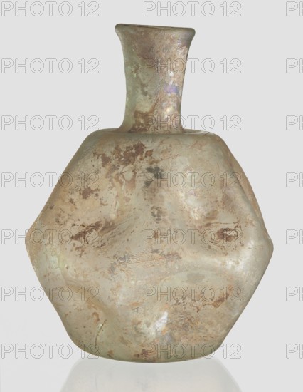Flask, unknown eastern workshop, 3rd–4th century AD, vessel, height 11 cm, A roughly hexagonal, bulbous vessel with a short flared neck and narrow mouth sits against a pale background; the surface shows a weathered, iridescent patina with colors including muted green, beige, brown, and hints of purple and gold, and it has mottled patches, irregular spotting, and a slightly pitted texture that gives it an aged, uneven appearance.