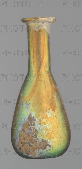 Flask, unknown eastern workshop, Goluchów Greater Poland Voivodeship, glass, antiquity, Roman Empire. Flask with a slender, pear-shaped body and a lower neck ending in a ring-shaped rim. Flat bottom. Covered with multicolored patina. 1st–2nd century, vessel, height 9.4 cm, A slender bottle with a narrow neck and a flared rim sits against a plain gray background, its surface showing an iridescent sheen of gold and green with areas of pale purple and silvery tones, along with patches of rougher, matte brown and beige where the finish appears worn; the overall shape tapers from a rounded base up to the elongated neck, and the rim shows some chips and uneven edges.