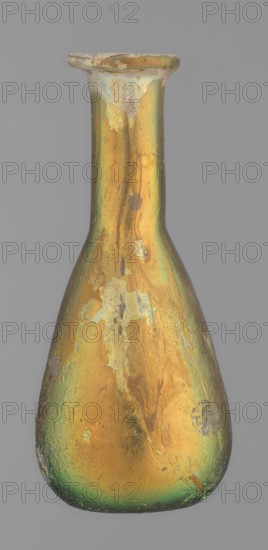 Flask, unknown eastern workshop, Goluchów Greater Poland Voivodeship, glass, antiquity, Roman Empire. Flask with a slender, pear-shaped body and a lower neck ending in a ring-shaped rim. Flat bottom. Covered with multicolored patina. 1st–2nd century, vessel, height 9.4 cm, A narrow-necked bottle with a flared rim shown against a plain gray background, the bottle surface displaying an iridescent sheen in gold and green tones with patches of duller, flaked areas and small rough spots, the body broadening toward the base and tapering to the neck, the rim appearing slightly chipped with uneven edges, and the overall texture looking worn with subtle variations of yellowish-gold, green, and muted brown on the surface.