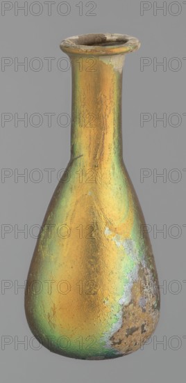 Flask, unknown eastern workshop, glass, antiquity, Roman Empire. Flask with a slender, pear-shaped body and a lower neck ending in a ring-shaped rim. Flat bottom. Covered with multicolored patina. 1st–2nd century, unknown place of origin; vessel, height 9.4 cm., A tall, narrow-necked vessel with a flared rim and a bulbous body, showing a textured iridescent surface with dominant gold and green tones and areas of brown and gray patina; the rim and parts of the neck have worn and chipped edges revealing a duller, rougher material, and there are patches of mottled discoloration and surface pitting near the base and on one side.