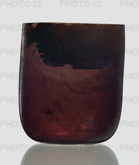 Bottle, unknown eastern workshop, 3rd century, vessel, height 4 cm, width 3.4 cm, A close-up of a rounded, translucent container with a smooth curved bottom resting on a reflective surface, showing a mix of deep burgundy and dark maroon tones with areas of darker almost black shading near the top, subtle patches of lighter reddish highlights, and some cloudy mottled textures and tiny surface imperfections visible through the material.