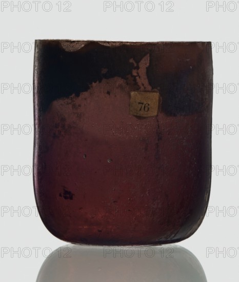 Bottle, unknown eastern workshop, Goluchów Greater Poland Voivodeship, 3rd century, vessel, height 4 cm, width 3.4 cm, A close-up of a rounded rectangular glass vessel with a smooth curved base, showing a translucent deep reddish-brown color that becomes darker near the top and edges, with areas of darker brown and almost black mottling and some lighter brown patches; the surface has small bubbles, pitting, and irregular texture, and there is a small aged paper-like square adhered near the upper center in a faded beige tone; the vessel sits on a reflective pale surface that shows a soft mirrored reflection, and the background is a very light, neutral color.