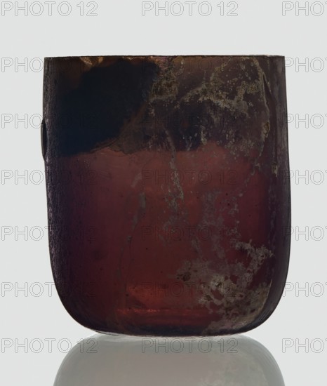 Bottle, unknown eastern workshop, Goluchów Greater Poland Voivodeship, 3rd century, vessel, height 4 cm, width 3.4 cm, A rounded, translucent object with a flat top and curved bottom sits against a pale background, showing a worn surface with mottled patches and irregular textures; its dominant colors are deep burgundy and dark brown with areas of black near the top and subtle hints of gray and muted rust, and a faint reflective sheen appears beneath it.
