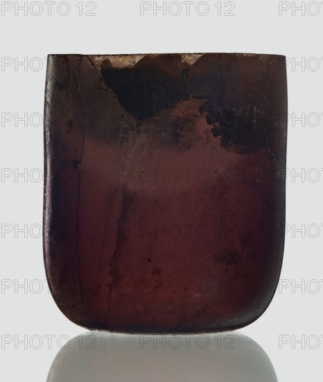 Bottle, unknown eastern workshop, Goluchów Greater Poland Voivodeship, 3rd century, vessel, height 4 cm, width 3.4 cm, A close-up of a rounded rectangular translucent object with a smooth, worn surface showing a mix of deep burgundy, dark brown, and near-black tones, with a few lighter reddish and amber patches near the top; the object rests on a reflective pale surface that produces a faint mirror image, and the background is a pale neutral gray.