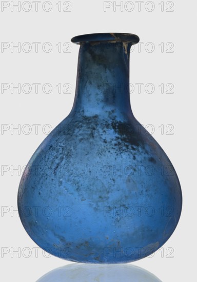Bottle, unknown eastern workshop, 1st–2nd century, vessel, height 7.5 cm, width 5.5 cm, A rounded glass vessel with a narrow neck and flared rim, predominantly blue with areas of darker blue and blackish mottling and some lighter blue patches, showing a matte, weathered surface texture with small blemishes and spots; the rim has a darker edge with a small lighter chip, and the object sits on a reflective pale surface against a light gray background.