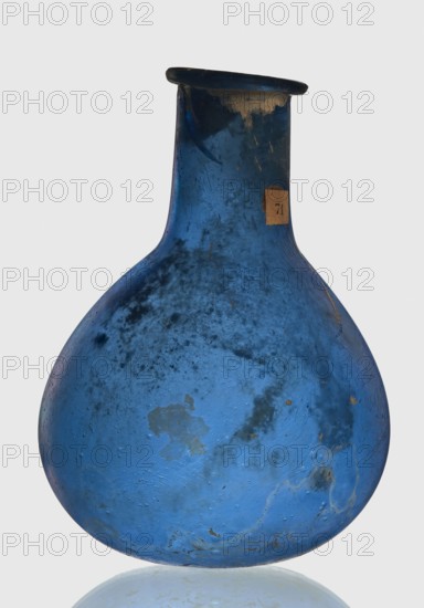 Bottle, unknown eastern workshop, Goluchów Greater Poland Voivodeship, 1st–2nd century, vessel, height 7.5 cm, width 5.5 cm, A single rounded glass vessel with a narrow neck and flared lip, shown against a pale background. The vessel is predominantly blue with areas of darker blue and some lighter blue patches. There are several mottled and cloudy dark spots and stains across the surface, along with a few small areas where the color appears worn or discolored to a tan or beige. The glass surface shows texture with tiny pits and irregularities and a few faint scratches. The overall shape is bulbous at the base tapering to a slim neck with a slightly uneven rim. The bottom edge sits on a reflective surface that produces a pale blue reflection.