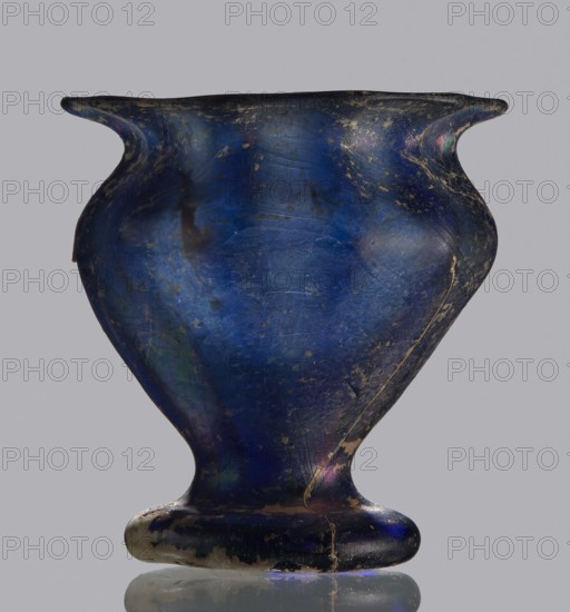 Krateriskos, unknown eastern workshop, Goluchów Greater Poland Voivodeship, 3rd–4th century AD, vessel, height 3.4 cm, width 3 cm, A single vessel with a rounded body and a flared rim sits centered against a plain background; the surface shows an iridescent deep blue as the dominant color with areas of darker navy and hints of purple, green, and subtle gold-toned weathering; the form narrows toward a short pedestal foot that is slightly flattened, and the exterior surface displays scratches, worn patches, and thin hairline lines following the curve of the body; the rim flares outward with gently upturned lips on opposite sides, and the overall finish appears glossy where light reflects, contrasting with matte worn spots.