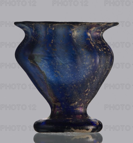 Krateriskos, unknown eastern workshop, Goluchów Greater Poland Voivodeship, 3rd–4th century AD, vessel, height 3.4 cm, width 3 cm, A single small vessel with a bulbous, curving profile and a flared rim, shown against a plain neutral background; the surface is a deep iridescent blue with areas of darker navy and hints of purple and green, and there are scattered worn patches and rougher spots where the finish is lighter and more matte, appearing beige or tan; the vessel sits on a rounded foot that reflects the same blue and dark tones, and the overall texture shows streaks and subtle scratches across the body.