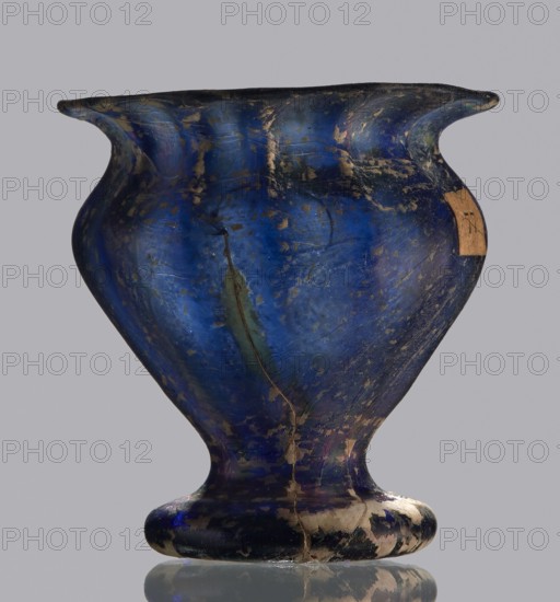 Krateriskos, unknown eastern workshop, Goluchów Greater Poland Voivodeship, 3rd–4th century AD, vessel, height 3.4 cm, width 3 cm, A rounded vessel with a flared rim and a short pedestal base, shown against a plain gray background, with a glossy surface in deep blue and patches of darker blue and black, along with areas of greenish discoloration and worn beige or tan spots; the surface shows scratches and irregular patches of missing material, and a small rectangular beige fragment attached near the rim; the vessel casts a faint reflection on the surface beneath it.