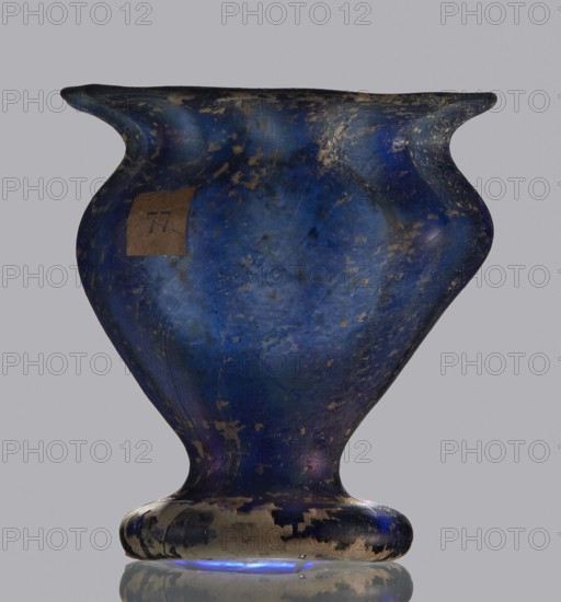 Krateriskos, unknown eastern workshop, Goluchów Greater Poland Voivodeship — 3rd–4th century AD, vessel, height 3.4 cm, width 3 cm, A single rounded vessel with a flared rim and a bulbous body sits against a plain light gray background, showing a worn surface with patches of darker and lighter coloration. The vessel is predominantly deep blue with areas of lighter blue and small spots of gold or tan wear scattered across its surface, and there are a few darker near-black areas along the rim and base. A small patch of brown paper is attached to one side of the body. The base shows a mix of blue, tan, and black wear and reflects faintly on the surface beneath it, producing a subtle blue glow at the very bottom.