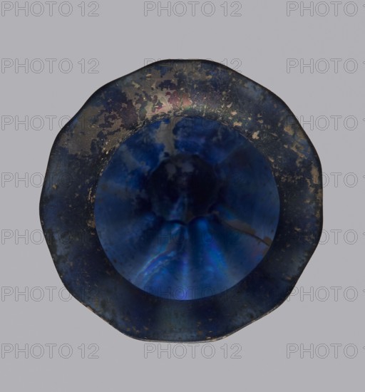 Krateriskos, unknown eastern workshop, Goluchów Greater Poland Voivodeship — 3rd–4th century AD, vessel, height 3.4 cm, width 3 cm, A circular object with an irregular rim is centered against a light gray background; the object shows a deep blue central area with iridescent shifts of lighter blue and purple, surrounded by a darker, mottled ring that includes shades of dark blue, black, brown, and hints of green and gold.