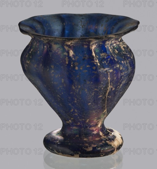 Krateriskos, unknown eastern workshop, Goluchów Greater Poland Voivodeship — 3rd–4th century AD, vessel; height 3.4 cm, width 3 cm, An iridescent vessel with a flared rim and a rounded body sitting on a short pedestal base, showing a worn surface with patches of loss; the dominant color is deep blue with visible areas of purple, green, gold, and black, and a few lighter beige areas where the surface is abraded.