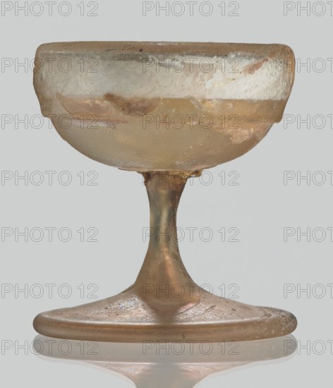Cup, Egyptian workshop, Goluchów Greater Poland Voivodeship, 3rd–4th century AD, Roman Empire 27 BC–476, vessel, height 3.1 cm, width 3.2 cm, A small translucent vessel with a rounded bowl and a flared base sits against a pale gray background; the glass shows a mix of pale amber, light brown, and hints of green with areas of opalescent sheen and cloudy white patches, surface irregularities, and roughened edges; the stem is slender and slightly tapered connecting the bowl to a broad flat foot that has a reflective underside and subtle iridescent speckles in pink and purple tones.