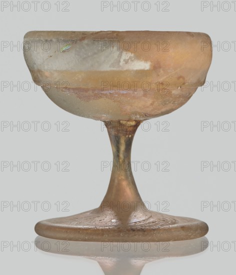 Cup, Egyptian workshop, Goluchów Greater Poland Voivodeship, 3rd–4th century AD, Roman Empire 27 BC–476, vessel, height 3.1 cm, width 3.2 cm, The image shows a single goblet-like object with a rounded bowl, a narrow stem, and a flat circular base, all appearing to be made of translucent material with a weathered surface. The object displays a range of colors including amber, pale gold, pinkish-beige, gray, and hints of green iridescence on the bowl. The surface shows areas of cloudiness, pitting, and uneven texture, with some glossy and some matte patches. The background is a plain light gray, and the object casts a faint reflection on a smooth reflective surface beneath it.