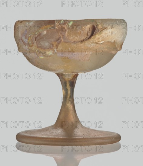 Cup, Egyptian workshop, Goluchów Greater Poland Voivodeship, 3rd–4th century AD, Roman Empire 27 BC–476, vessel, height 3.1 cm, width 3.2 cm, A translucent goblet with a wide shallow bowl and a tapered stem standing on a broad rounded foot, showing irregular surface texture and areas of roughness and flaking. The object displays a palette of beige, tan, amber, and pale brown with iridescent highlights in green and purple on parts of the bowl, along with patches of lighter cream and off-white. The stem and foot have a muted golden-brown tone with slightly darker brown streaks, and the base reflects faintly on the surface beneath.