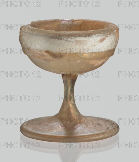 Cup, Egyptian workshop, Goluchów Greater Poland Voivodeship — 3rd–4th century AD, Roman Empire 27 BC–476, vessel; height 3.1 cm, width 3.2 cm, A small translucent goblet with an iridescent sheen, showing a pale amber and gold color on the bowl and stem, with areas of whitish, chalky residue on the upper rim; the stem is slender and tapers into a round flattened base that reflects soft hints of pink and green, and the surface shows a slightly rough, weathered texture against a neutral gray background.