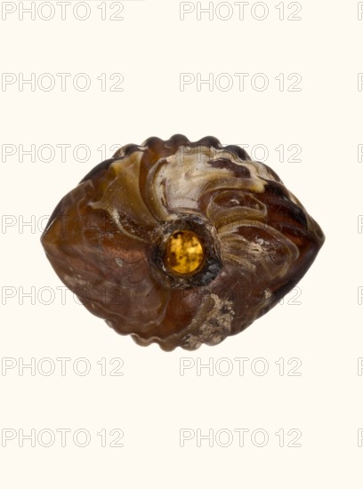 Balsamarium in the shape of a double shell, Goluchów Greater Poland Voivodeship, 2nd–3rd century AD, vessel, height 7.1 cm, An irregular, oval-shaped object resembling a carved bead sits against a plain light background; its surface shows a mix of amber, brown, yellow, cream, and dark brown tones with glossy and matte areas, wavy carved ridges and grooves across the top and bottom, a central round hole revealing a golden-yellow inner surface, and patches of lighter deposits or residue on parts of the exterior.