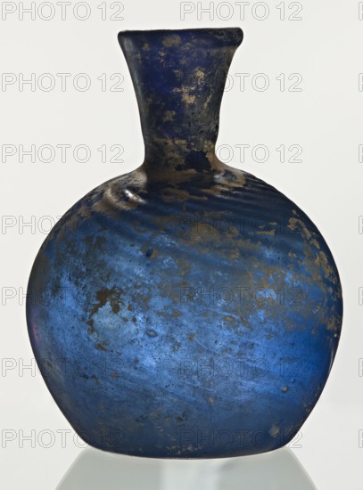 Bottle, unknown eastern workshop, Goluchów Greater Poland Voivodeship, 3rd–4th century, vessel, height 10 cm, width 3.8 cm, A rounded glass vessel with a short neck and flared rim, predominantly deep blue with irregular patches and speckles of darker tones and areas of brownish weathering, showing a mottled, uneven surface texture and subtle variations of lighter and darker blue across its bulbous body, photographed against a plain light background.
