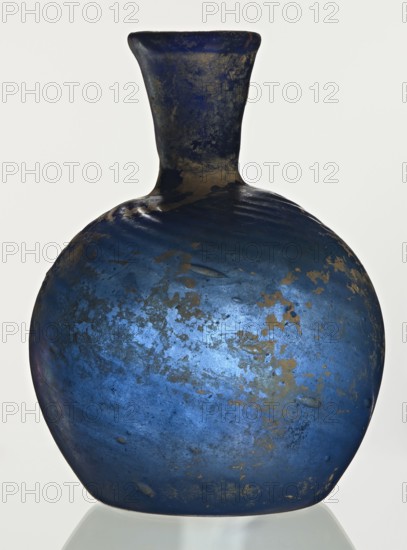 Bottle, unknown eastern workshop, Goluchów Greater Poland Voivodeship, 3rd–4th century, vessel, height 10 cm, width 3.8 cm, A rounded bottle with a narrow neck and flared rim, showing a worn, textured surface in deep blue with patches of metallic gold and darker blue mottling; the finish has irregular spots and small indentations, and the base reflects faintly on a pale surface.
