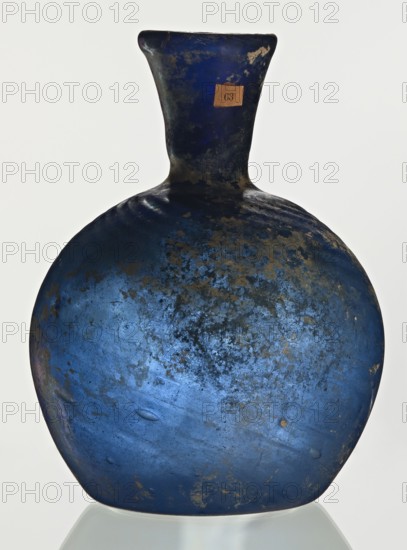 Bottle, unknown eastern workshop, Goluchów Greater Poland Voivodeship, 3rd–4th century, vessel, height 10 cm, width 3.8 cm, A worn rounded vessel with a narrow neck and flared rim, shown against a light background; the surface is predominantly deep blue with areas of darker navy and patches of lighter blue, along with several irregular mottled spots of brown and tan discoloration and some textured rough areas; the rim and neck show darker blue and near-black tones; the base sits on a reflective surface that shows a pale gray reflection.