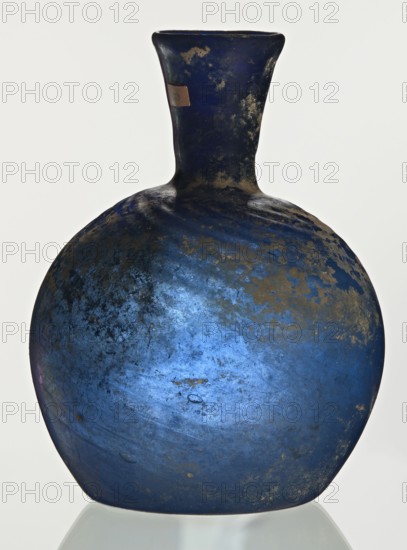 Bottle, unknown eastern workshop, 3rd–4th century, vessel, height 10 cm, width 3.8 cm, A rounded vessel with a narrow neck and flared rim, shown against a pale background, featuring a dominant deep blue color with areas of lighter blue and patches of dark brownish-gold weathered texture across its surface; the neck appears darker blue with spots of the same brownish-gold, and the vessel sits on a reflective surface that mirrors a faint pale grayish reflection.