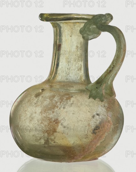 Jug, Egyptian workshop, spherical flattened body, cylindrical neck, flared spout and ribbon-shaped handle with thickened edges, 6th–4th century BC, vessel, height 7 cm, width 6 cm, A translucent glass jug with a rounded body and a narrow neck flaring into a wide rim, showing a weathered, iridescent surface in pale green and yellow tones with areas of brownish and pinkish discoloration, and a curved greenish handle attached near the rim and shoulder with a decorative leaf-like terminal, all set against a plain light background.