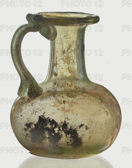 Jug, spherical flattened body, cylindrical neck, flared spout and ribbon-shaped handle with thickened edges; Egyptian workshop; 6th–4th century BC, unknown place of origin; vessel; height 7 cm, width 6 cm., A translucent glass jug with a rounded body, narrow neck, and flared rim, showing a smooth, curved handle attached from rim to shoulder; the glass surface displays a mix of pale green, yellowish, and amber tones with areas of iridescent sheen and cloudy, weathered patches; there are several dark mottled spots on the lower body and scattered flecks and abrasions across the neck and shoulder; overall the object appears worn with a slightly roughened texture and subtle variations in color intensity.