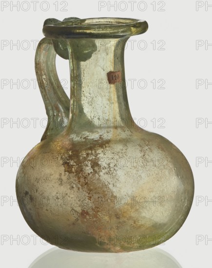 Jug, spherical flattened body, cylindrical neck, flared spout and ribbon-shaped handle with thickened edges, Egyptian workshop, 6th–4th century BC, vessel, height 7 cm, width 6 cm, A glass jug with a rounded body, a narrow neck, a flared rim, and a single handle; the glass appears translucent with a greenish tint, showing areas of pale green, yellow-green, and muted brownish discoloration and patina, along with patches of iridescent sheen and cloudy whitish deposits; the handle is attached from rim to shoulder and shares the greenish color with some darker streaks; the surface shows weathered texture with rough mottled areas and smooth glossy sections; the background is a plain light gray.