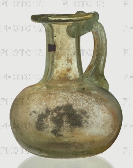 Jug, spherical, flattened body, cylindrical neck, flared spout and ribbon-shaped handle with thickened edges; Egyptian workshop; 6th–4th century BC ; vessel; height 7 cm, width 6 cm., A translucent glass jug with a rounded body, a narrow neck that flares at the rim, and a curved handle attached from the rim to the shoulder; the glass shows weathered iridescence and surface encrustation with areas of cloudy white, pale green, and muted gold tones, plus darker grayish patches on the body, and the background appears pale gray.