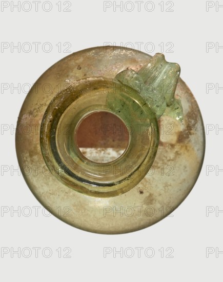 Jug, Egyptian workshop, spherical flattened body, cylindrical neck, flared spout and ribbon-shaped handle with thickened edges; 6th–4th century BC, unknown place of origin; vessel; height 7 cm, width 6 cm., A round glass object viewed from above with a central circular opening and a short cylindrical neck, showing a small handle-like protrusion on one side; the glass surface displays a mix of green and yellow tones with areas of pale brownish and off-white discoloration and some darker specks, and the background is a light gray.