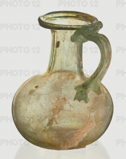 Jug, Egyptian workshop, spherical flattened body, cylindrical neck, flared spout and ribbon-shaped handle with thickened edges. 6th–4th century BC, origin unknown. Vessel; height 7 cm, width 6 cm., The image shows a translucent glass jug with a rounded body, a narrow neck, a flared rim, and a curved handle attached to the shoulder; the glass has an aged, iridescent surface with areas of cloudy opalescence and weathered spots. The visible colors include pale green, muted yellow-gold, hints of pinkish-orange, and some brownish discoloration, with reflective highlights that give slight metallic sheens. The handle is a greener tone and has a sculpted leaf-like attachment at its base, and the overall surface shows pitting, streaks, and worn textures consistent across the body and neck.