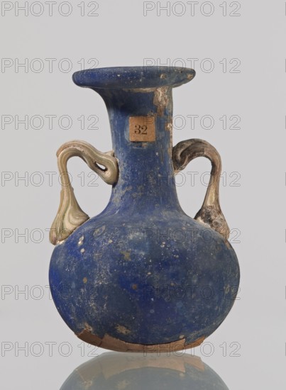 Amphora, bulging belly with a rounded bottom, long cylindrical neck with a thick ring-shaped rim; handles attached to the shoulders and halfway up the neck. 3rd–4th century, Roman Empire 27 BC–476. Vessel. Height 4.7 cm, width 3 cm., A rounded vessel with a narrow neck and flaring rim, showing a worn deep blue surface with lighter blue and pale spots and areas of abrasion, with two ornate handles that are pale beige and cream with some brown tones, and a strip of brownish material along the bottom edge; the background is a pale gray.