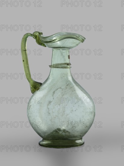 Oinochoe. Transparent, light green glass. Spherical belly on a foot with a clearance, created by pressing inward the central part of the bottom of the vessel; decoration on the cylindrical neck in the form of a single glass thread of the same color, wound horizontally. The handle is glued to the body and extends in an arc to the edge of the trefoil rim; free-blown. 4th century, Cyprus; vessel oinochoe. Base diameter 4.8 cm; weight 0.062 kg. Type: Isings form 124a., A transparent glass pitcher with a rounded body and a narrow neck topped by a flared, irregular rim and an undulating lip, featuring a curved handle attached to the rim and body; the glass shows a few internal bubbles and surface irregularities, with streaks and cloudy patches; colors visible are pale green and a slightly darker green on the handle and base, set against a neutral gray background.