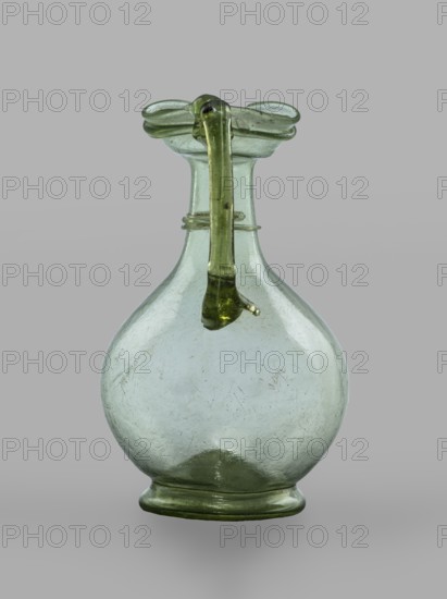 Oinochoe, Transparent, light green glass. Spherical belly on a foot with a clearance, created by pressing inward the central part of the bottom of the vessel; decoration on the cylindrical neck in the form of a single glass thread of the same color, wound horizontally. The handle is glued to the arms and extends in an arc to the edge of the trefoil rim; free-blown. 4th century, Cyprus; vessel oinochoe. Base diameter 4.8 cm, weight 0.062 kg. Type: Isings form 124a., A blown glass vessel with a rounded body and a narrow neck topped by a flared rim, featuring a looped handle that extends from the rim down toward the body; the glass appears translucent with green and pale green tones, and the surface shows visible scratches and small irregularities, while the background is a uniform light gray.